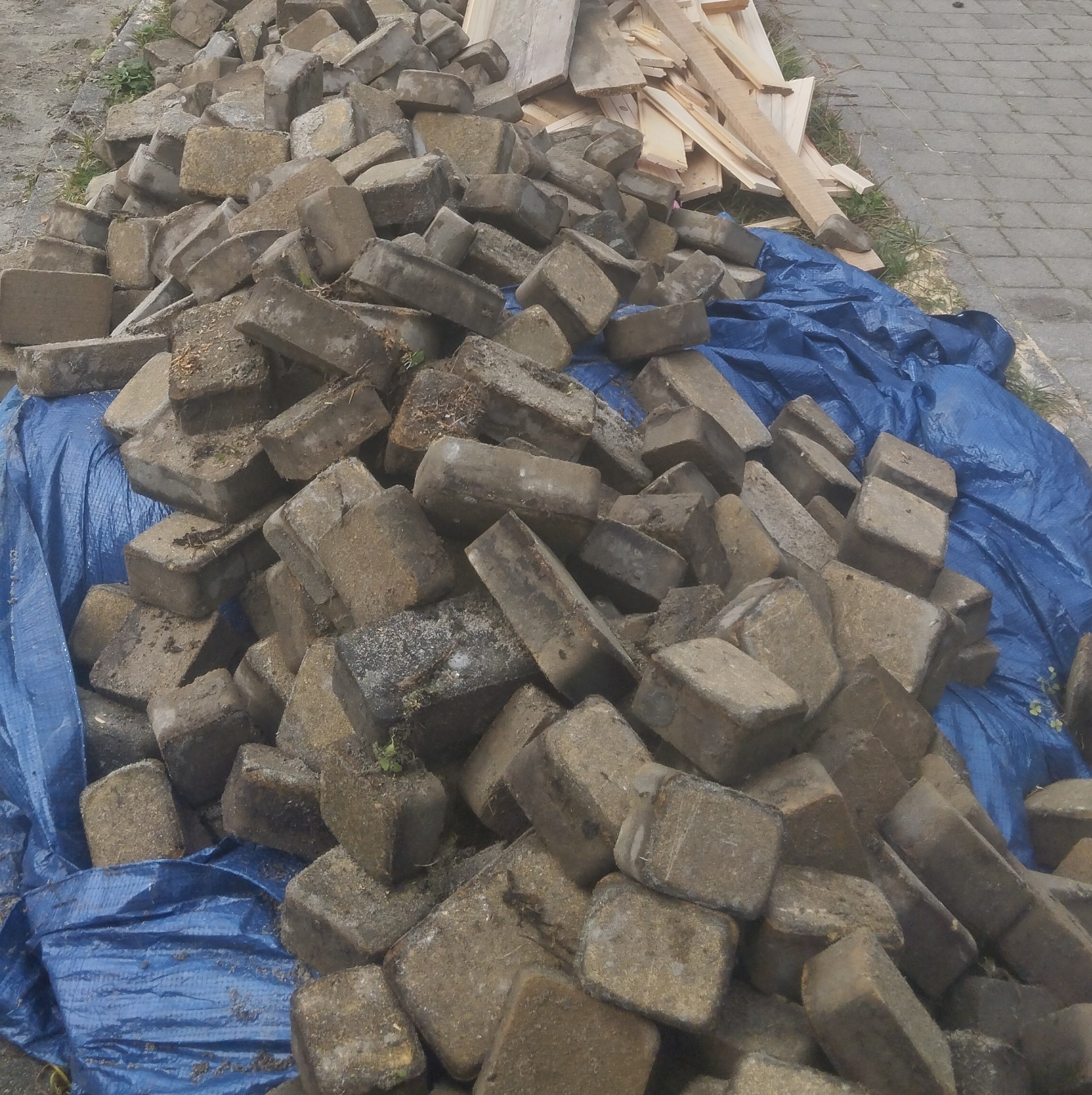 Stos szarych, betonowych kostek brukowych ułożonych na niebieskiej plandece, obok leżą drewniane deski