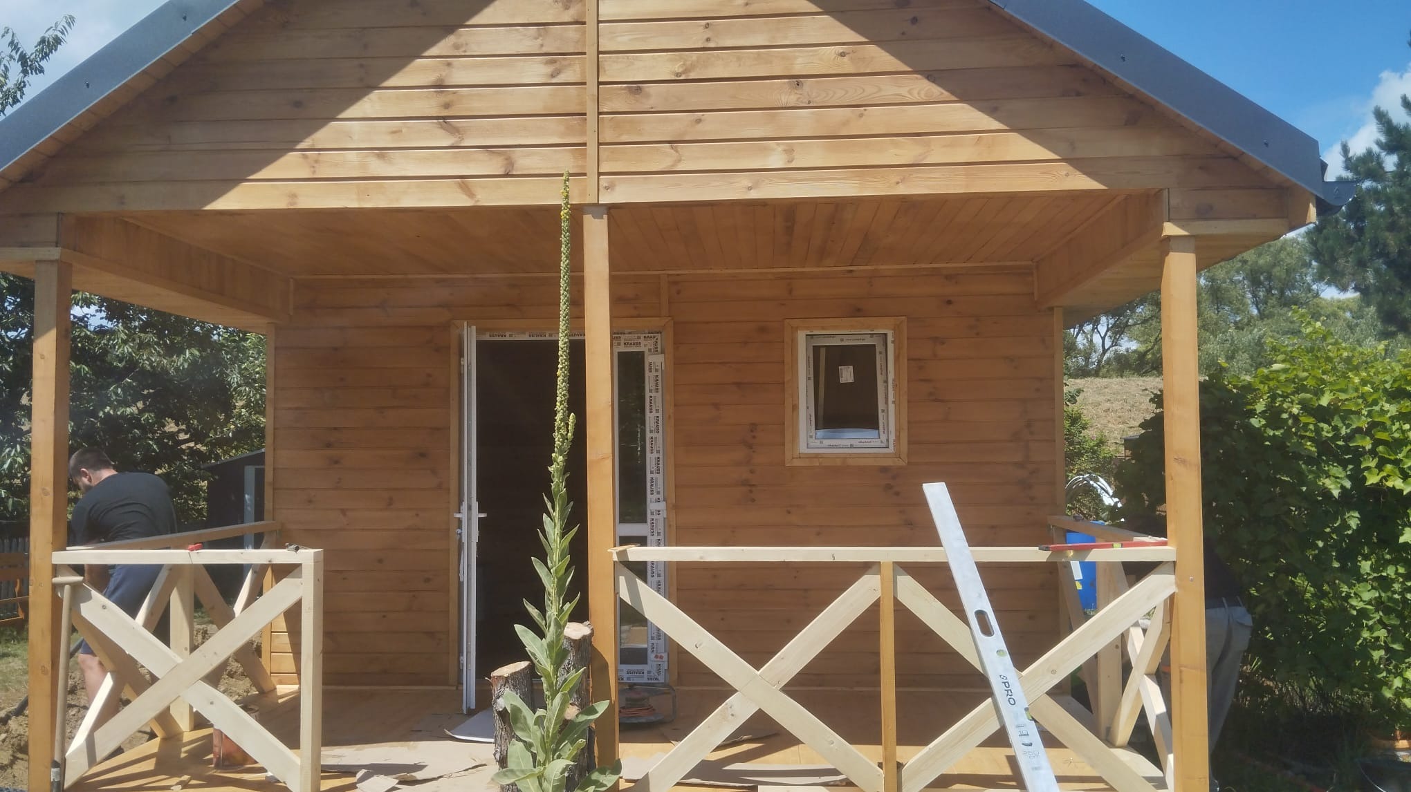 W trakcie budowy drewnianego domu szkieletowego z widocznym fragmentem elewacji, balustrady werandy w trakcie montażu, poziomica na balustradzie, wysoka roślina przed wejściem.