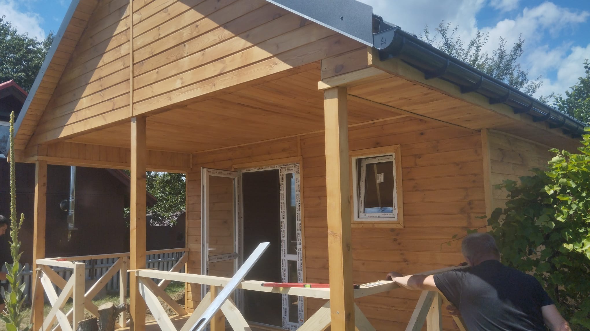 Drewniany domek letniskowy z zadaszonym tarasem w trakcie budowy, widoczne otwarte drzwi, okno z ramą PCV, czarne rynny i pracownik montujący balustradę tarasu.