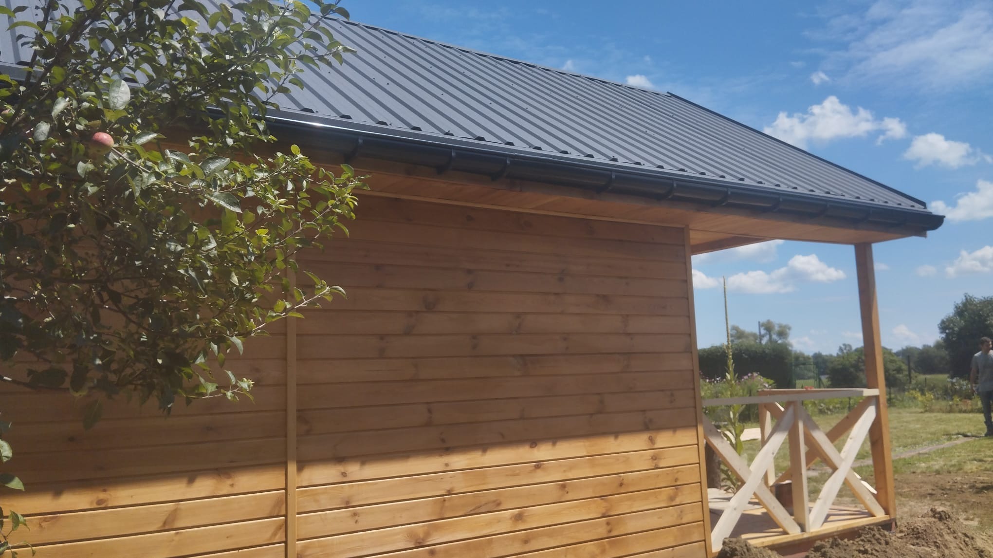 Fragment drewnianego domu z metalowym dachem, widoczny z boku, z elementami werandy i młodym drzewem owocowym w pierwszym planie.
