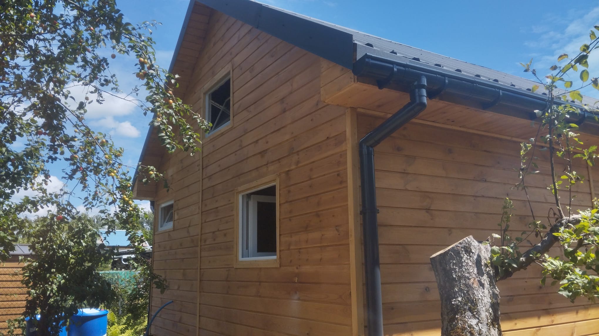 Zewnętrzny widok domu z drewnianą elewacją, czarnym dachem i rynnami, częściowo zasłoniętego przez drzewo owocowe i pień ściętego drzewa na pierwszym planie.