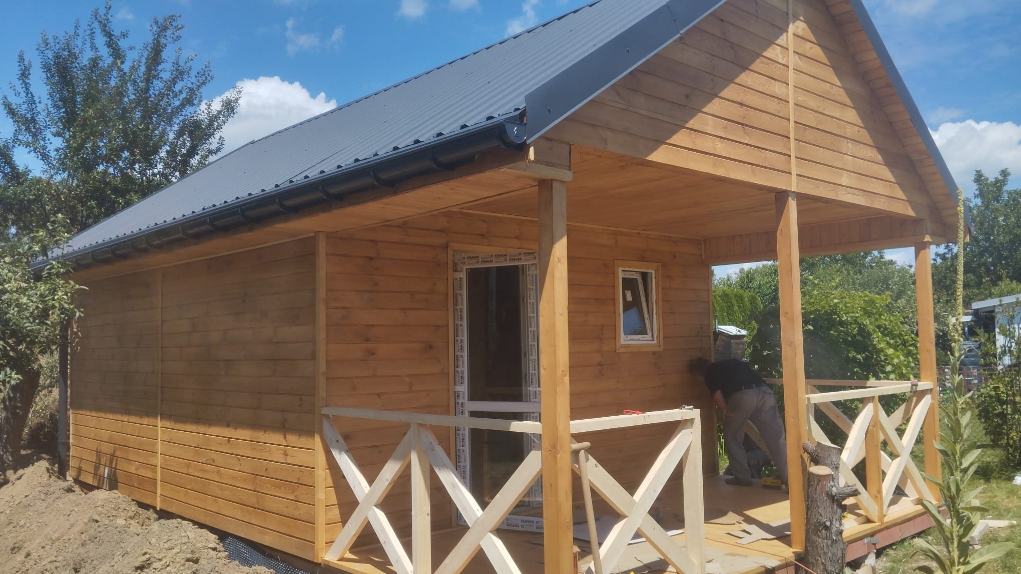 Niski, drewniany budynek z zadaszonym tarasem w trakcie budowy, widoczny pracownik montujący elementy wykończeniowe na zewnątrz, teren wokół budynku częściowo przygotowany.
