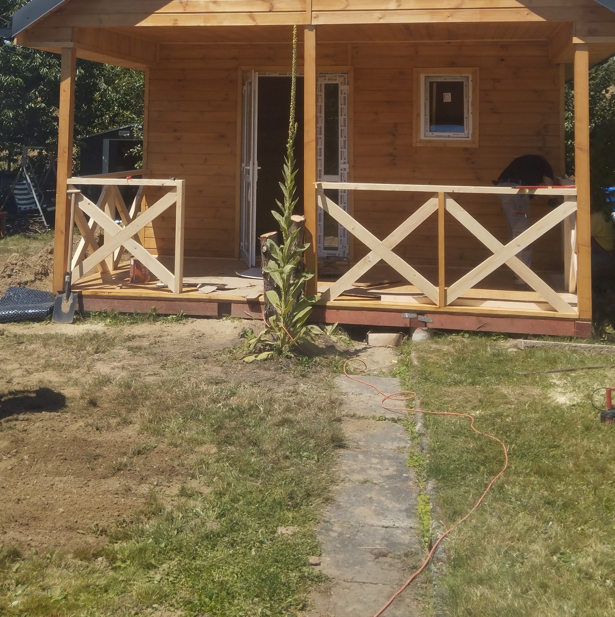 Drewniany domek letniskowy w trakcie budowy, widoczne elementy konstrukcyjne werandy z krzyżowymi belkami, narzędzia budowlane i osoba pracująca przy wykończeniu.