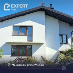 Dwukondygnacyjny dom jednorodzinny z białą elewacją i brązowymi ramami okiennymi, zwieńczony ciemnoszarym dachem z kominem i ozdobną metalową wiatrowskazem w kształcie samolotu, widziany z poziomu...