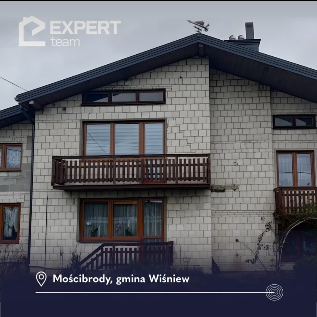 Niedokończony dwukondygnacyjny dom z białej cegły z drewnianymi oknami i balkonami, widoczna nieocieplona ściana szczytowa, na dachu wiatrowskaz w kształcie samolotu.