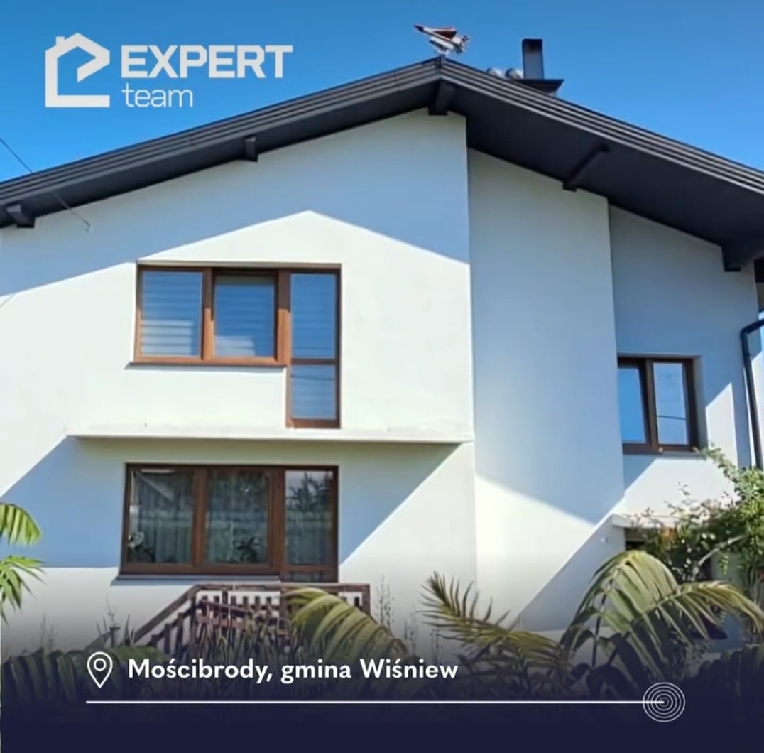 Dwukondygnacyjny dom jednorodzinny z białą elewacją i brązowymi ramami okiennymi, zwieńczony ciemnoszarym dachem z kominem i ozdobną metalową wiatrowskazem w kształcie samolotu, widziany z poziomu...