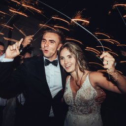 Krzysztof Kuźniar - Uśmiechnięta para młoda świętuje z gośćmi, trzymając zapalone zimne ognie, widoczne detale sukni ślubnej i garnituru.