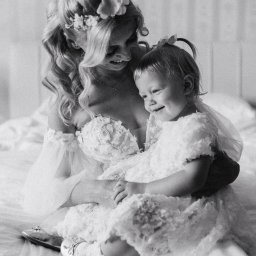 Krzysztof Kuźniar - Czarno-białe zdjęcie uśmiechniętej panny młodej w koronkowej sukni z kwiatami we włosach, trzymającej na kolanach uśmiechniętą małą dziewczynkę w podobnej koronkowej sukience i baletkach.