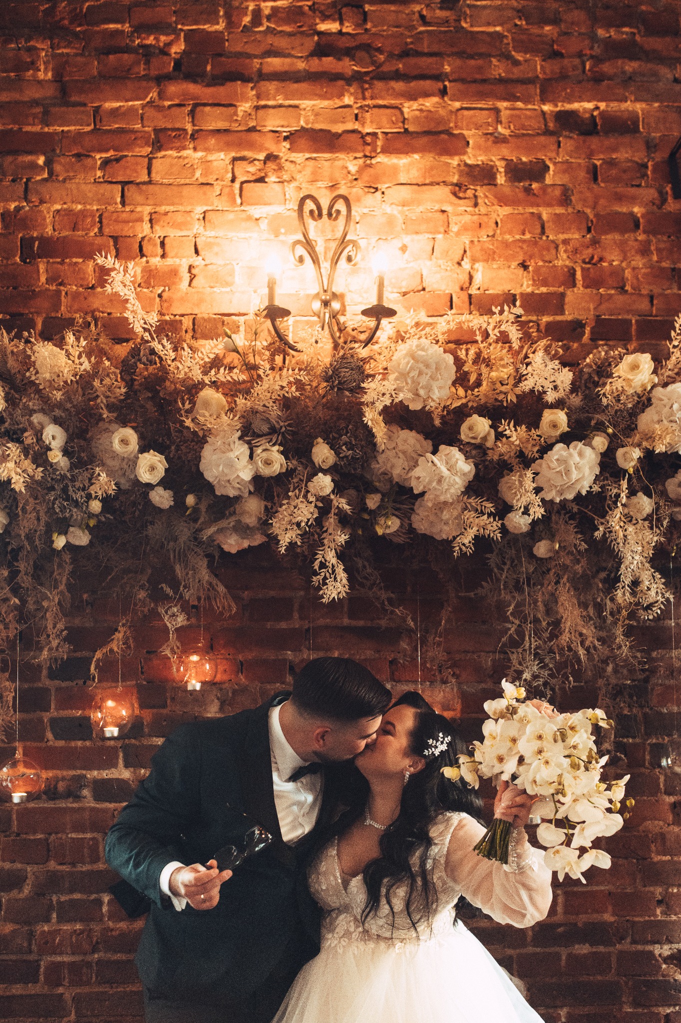 Pocałunek nowożeńców na tle ceglanej ściany z kwiatową dekoracją i kandelabrem, panna młoda trzyma bukiet białych orchidei.