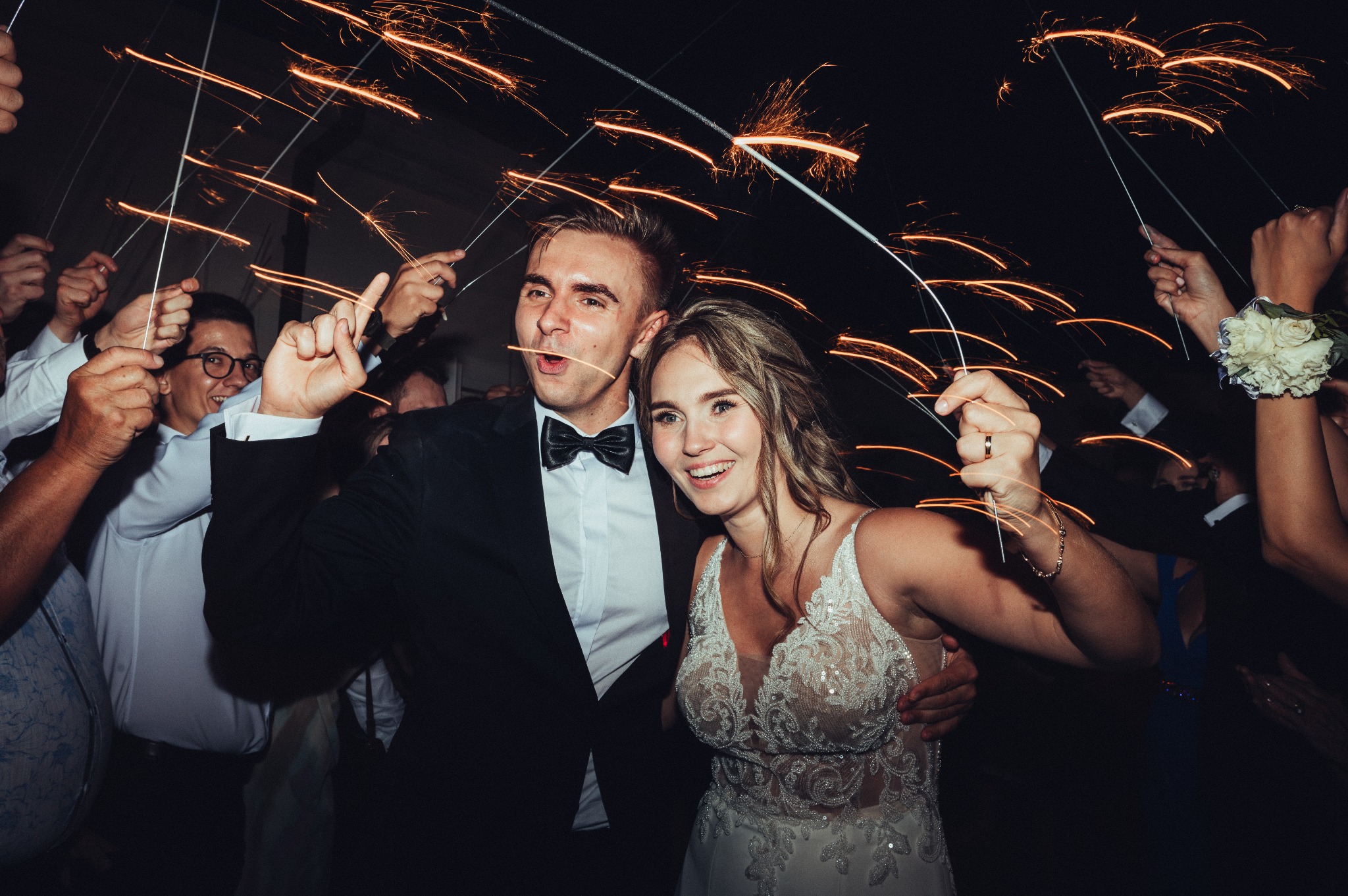 Uśmiechnięta para młoda świętuje z gośćmi, trzymając zapalone zimne ognie, widoczne detale sukni ślubnej i garnituru.