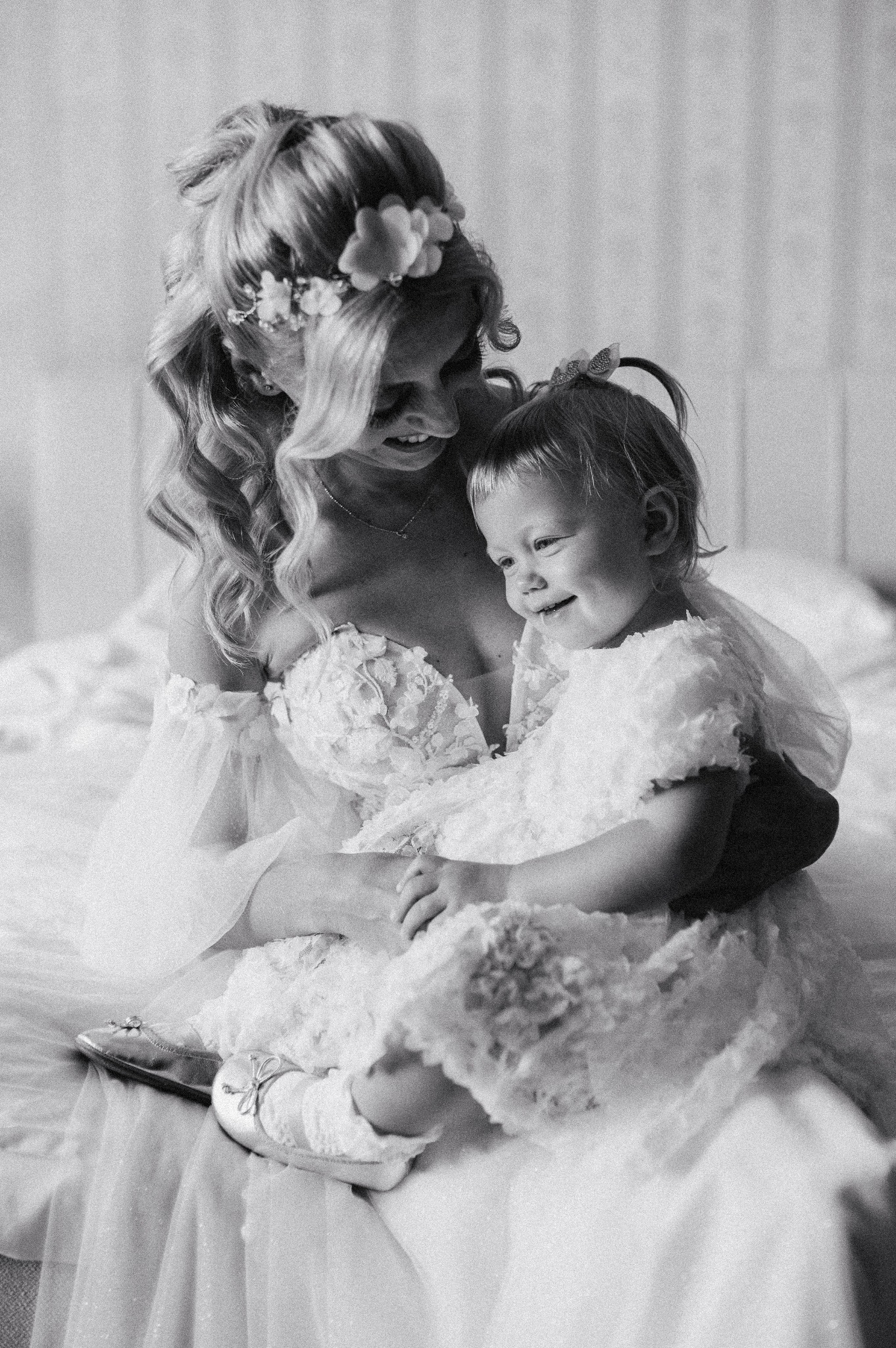Czarno-białe zdjęcie uśmiechniętej panny młodej w koronkowej sukni z kwiatami we włosach, trzymającej na kolanach uśmiechniętą małą dziewczynkę w podobnej koronkowej sukience i baletkach.