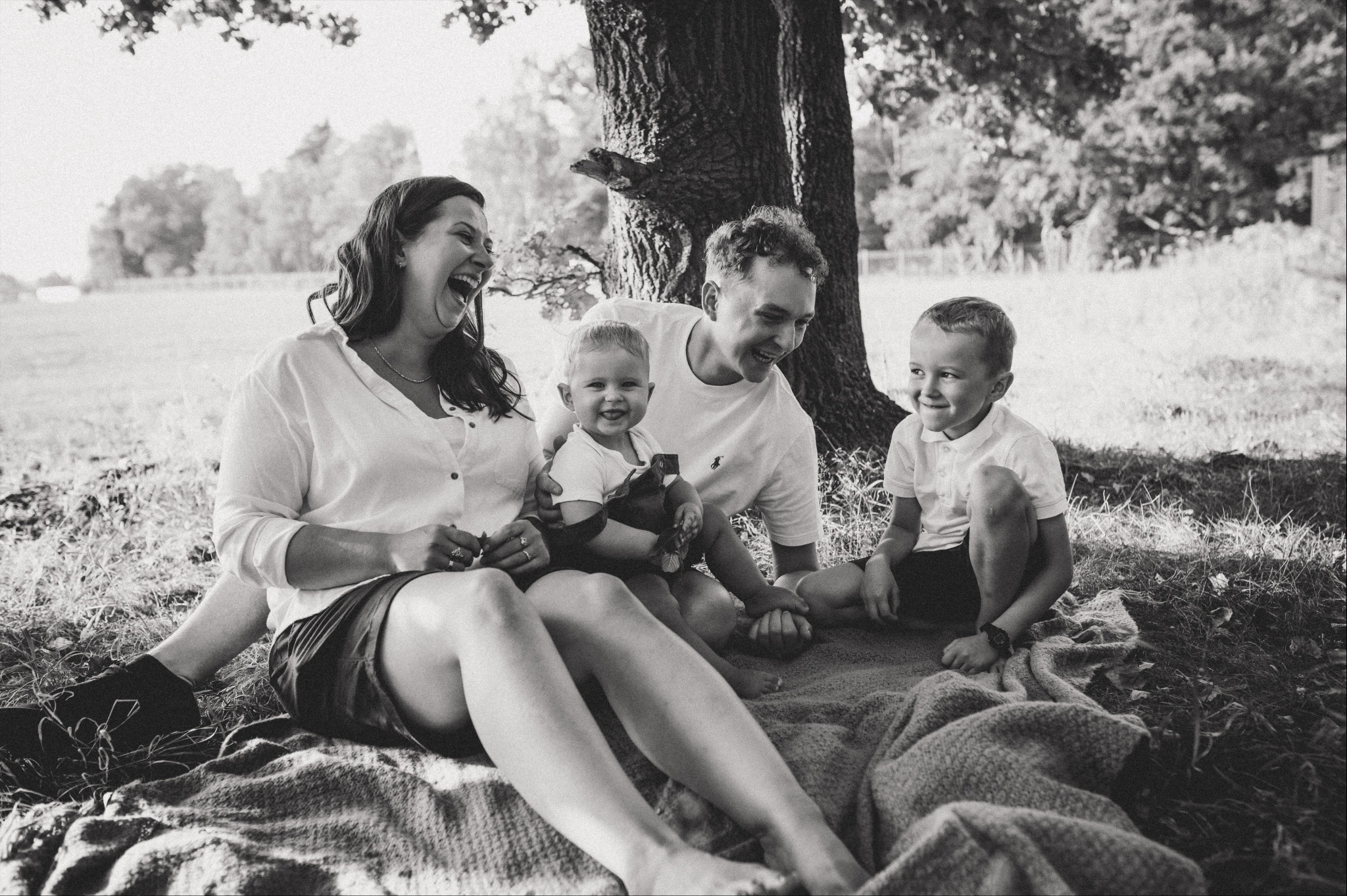 Czarno-białe zdjęcie uśmiechniętej rodziny siedzącej na kocu pod drzewem, z wyraźnym pierwszym planem trawy i liści.