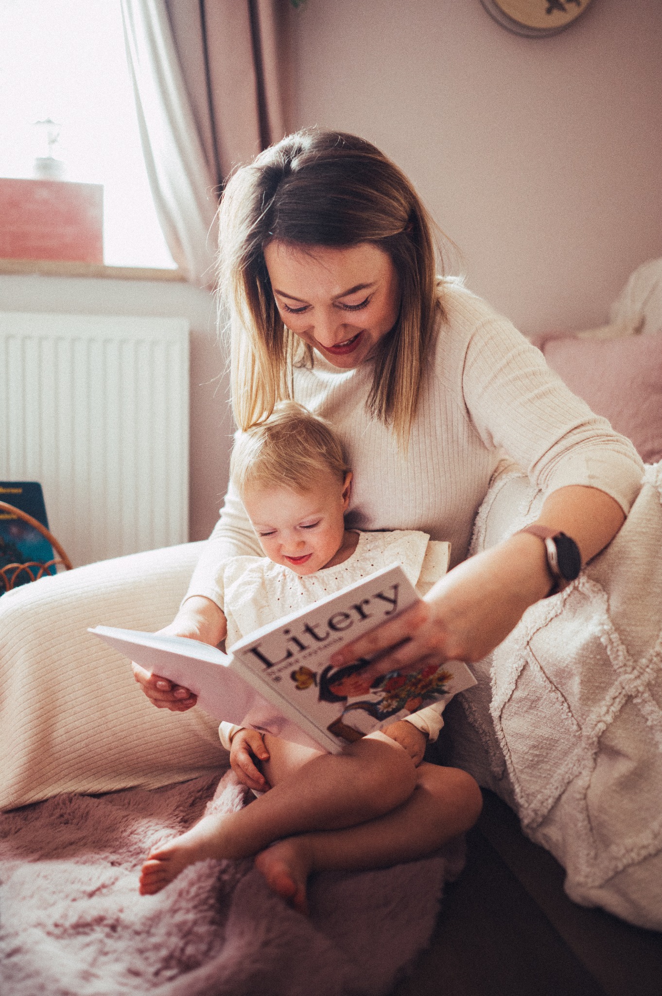 Matka z blond włosami czyta książkę 'Litery' córce na puszystym, różowym dywanie w pokoju z białym grzejnikiem.