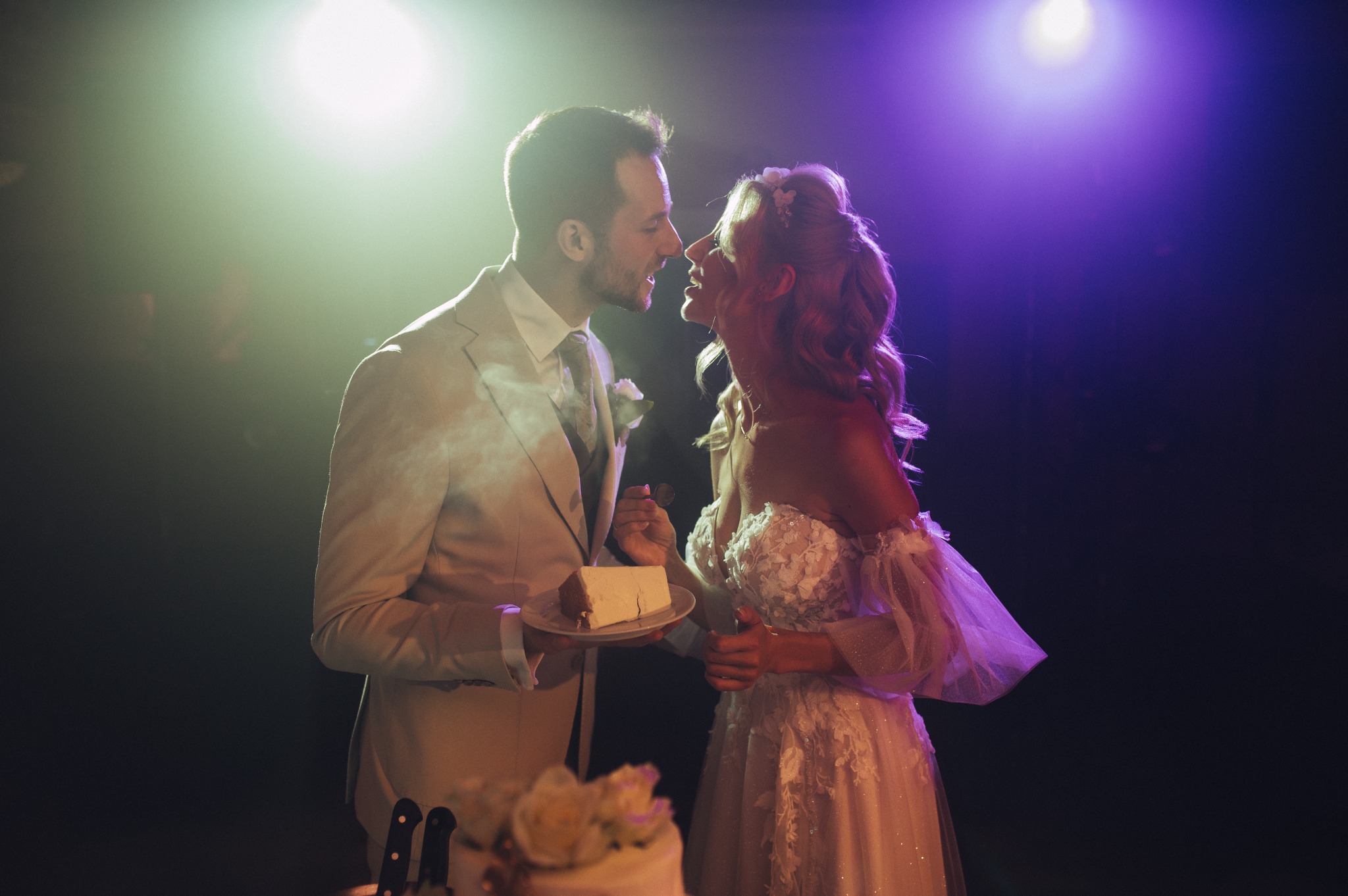 Nowożeńcy w trakcie krojenia tortu weselnego; panna młoda w białej sukni z koronkowymi aplikacjami, pan młody w beżowym garniturze podaje kawałek tortu na talerzyku.