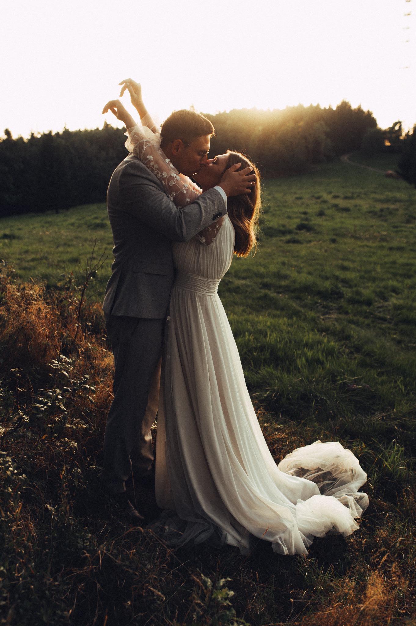 Para młoda w objęciach całuje się na tle zachodzącego słońca i łąki, panna młoda w zwiewnej sukni z koronkowymi rękawami, pan młody w szarym garniturze.