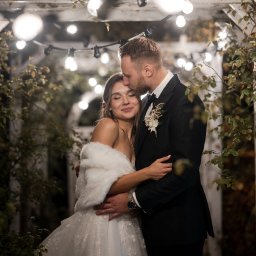 Mateusz Politański Herbaty z Nepalu - Elegancka para młoda w objęciach pod drewnianą pergolą z girlandami świetlnymi. Pan młody całuje pannę młodą w czoło. Romantyczna atmosfera.