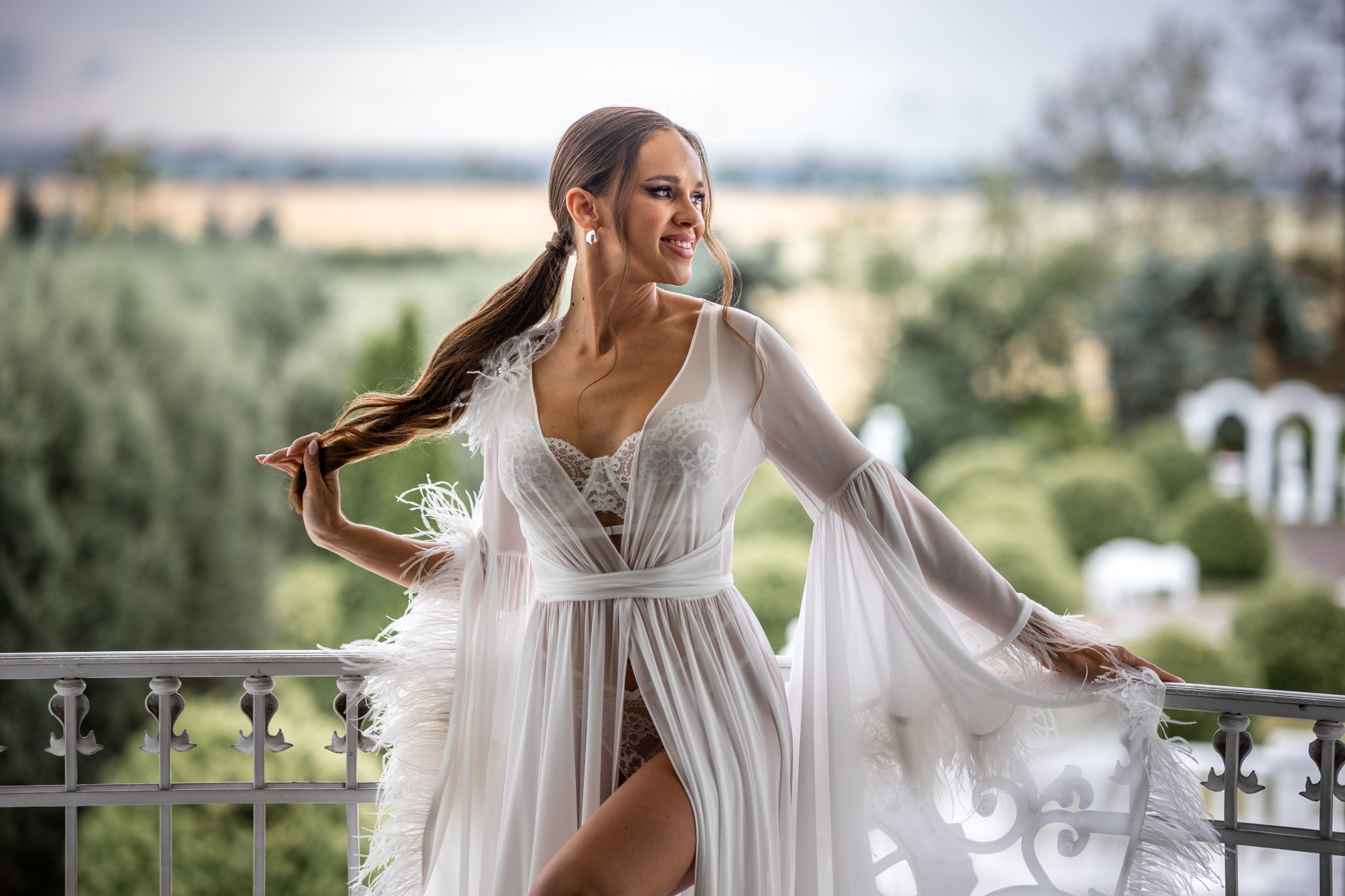 Uśmiechnięta panna młoda w białym, zwiewnym szlafroku z piórami, stojąca na balkonie z widokiem na zielony krajobraz, trzymająca kosmyk włosów.