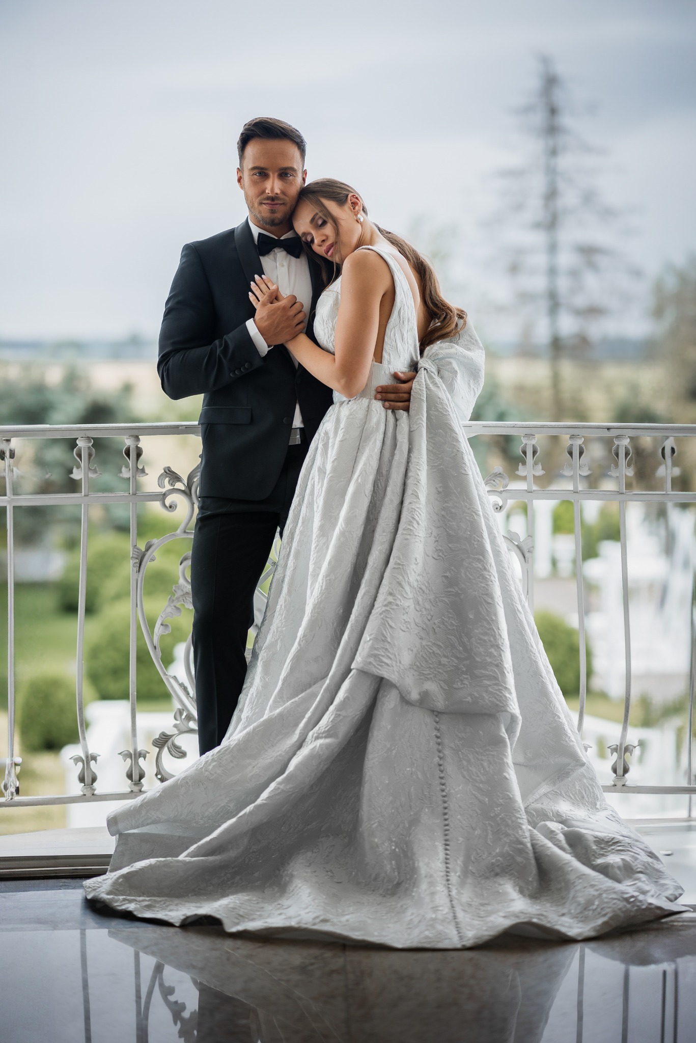 Elegancka para młoda: mężczyzna w czarnym smokingu i kobieta w srebrzystej sukni ślubnej z dużym kokardą, pozują na balkonie z ozdobną balustradą, w tle zieleń.