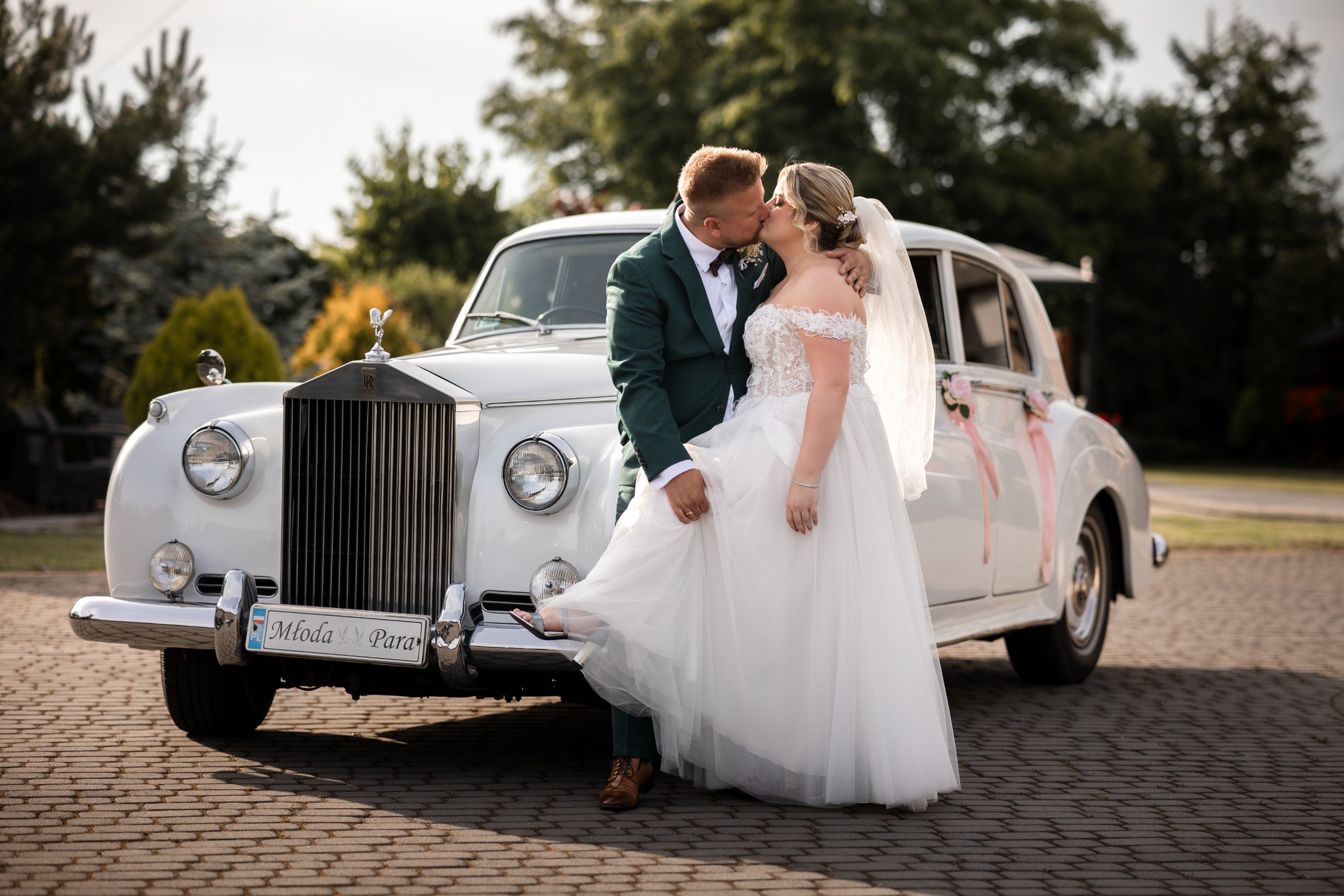 Pocałunek pary młodej na tle białego, zabytkowego Rolls Royce'a z dekoracjami kwiatowymi i tablicą 'Młoda Para', on w butelkowej zieleni garniturze, ona w koronkowej sukni ślubnej z odkrytymi...