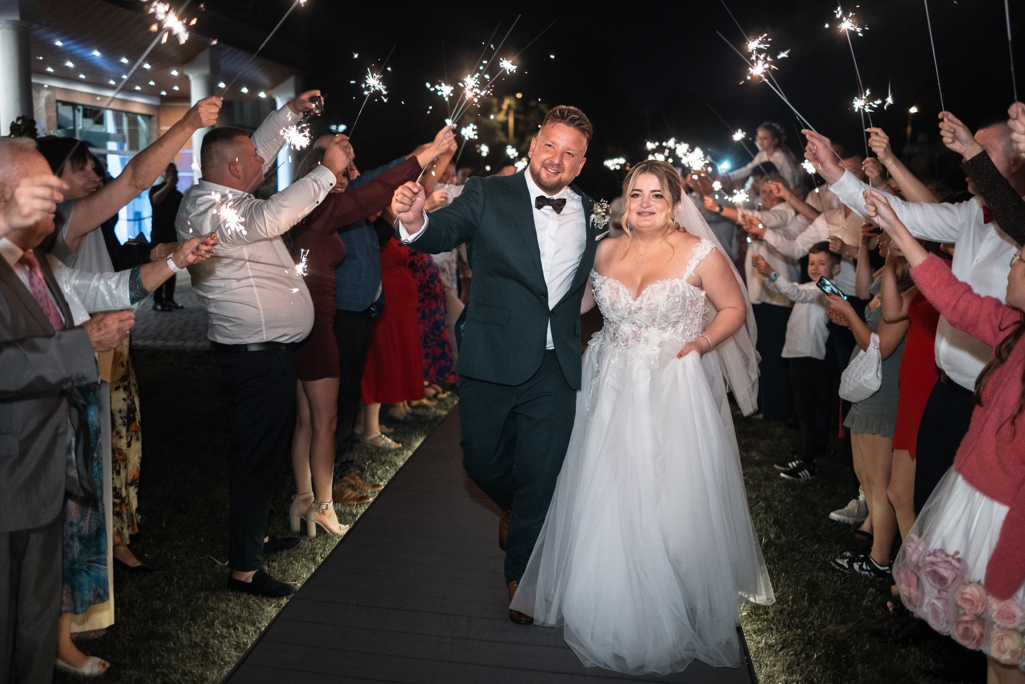 Para młoda przechodzi przez szpaler gości weselnych trzymających zapalone zimne ognie; panna młoda w białej sukni z koronkową górą, pan młody w ciemnozielonym garniturze z muszką.