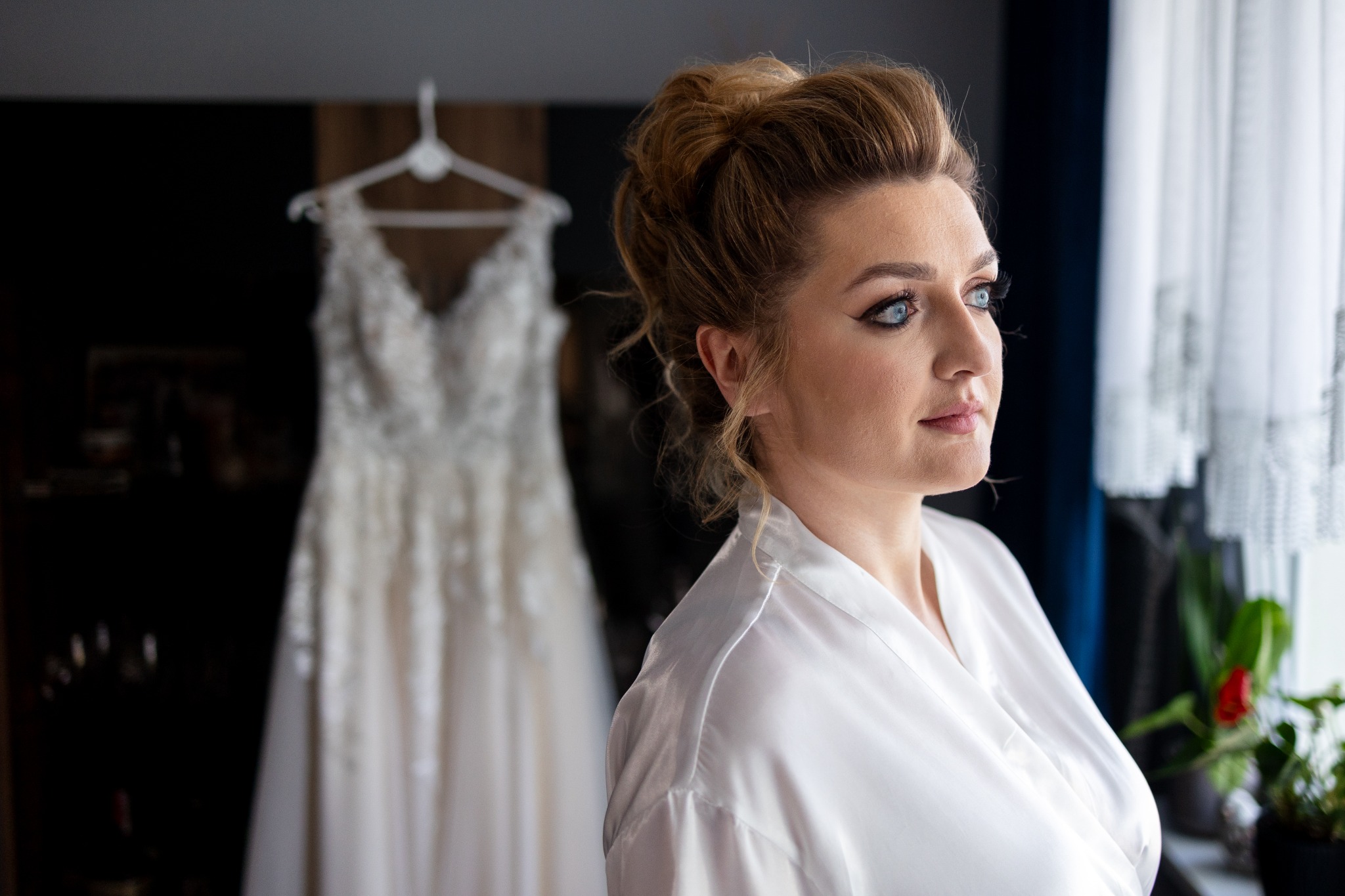 Panna młoda w szlafroku spogląda w okno, w tle suknia ślubna na wieszaku i firanka z wzorem.