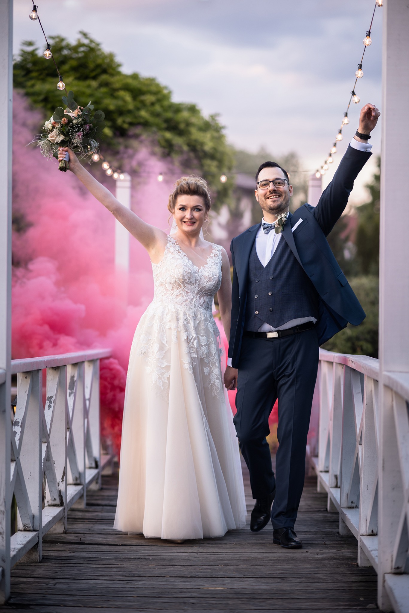 Uśmiechnięta para młoda na drewnianym moście z bukietem i uniesioną ręką na tle różowego dymu i girlandy światełek.