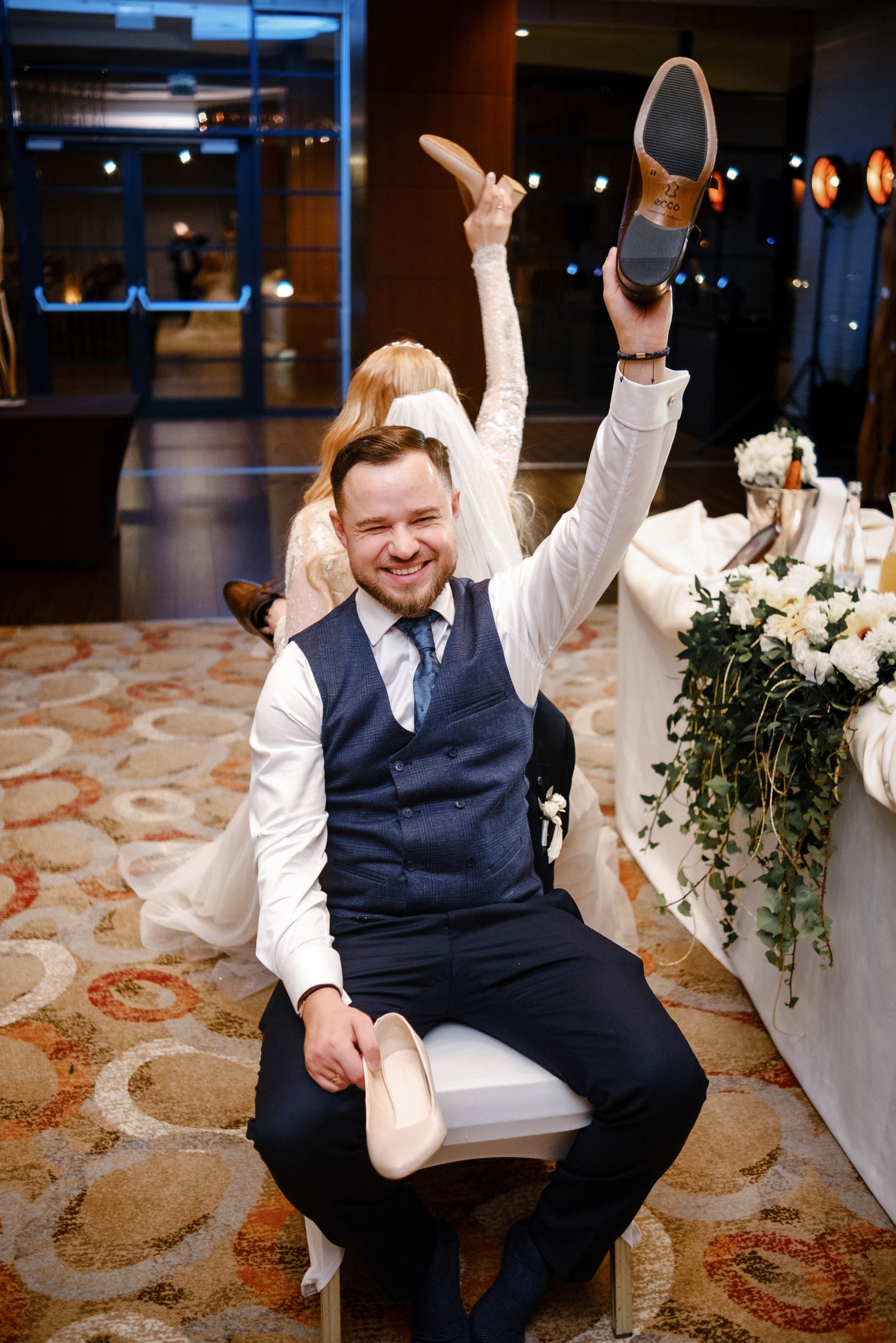 Uśmiechnięty pan młody siedzi na krześle, trzymając w jednej ręce pantofel damski, a w drugiej męski but podniesiony do góry, w tle panna młoda również z podniesionym butem.