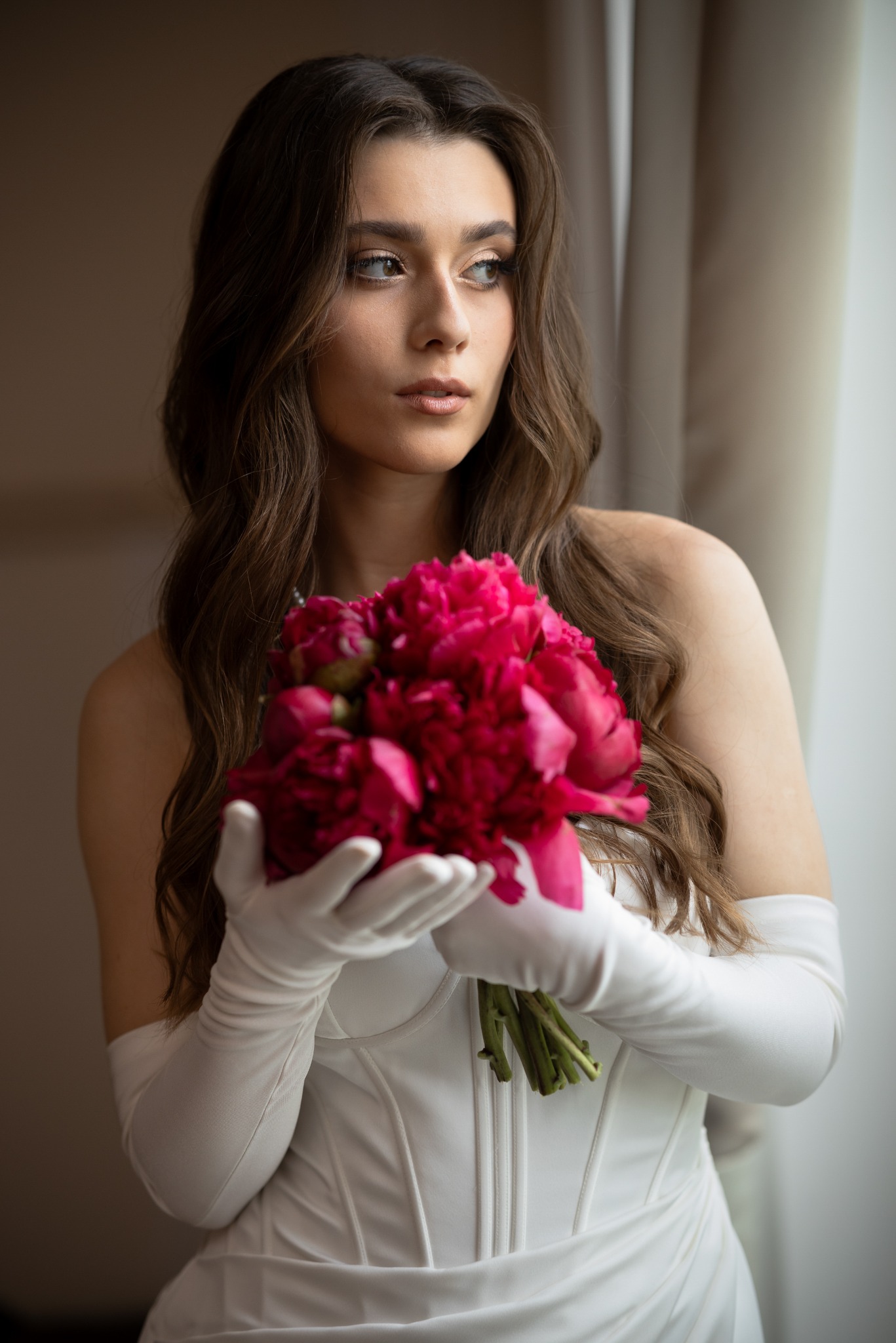 Panna młoda w białej sukni i długich rękawiczkach trzyma bukiet ciemnoróżowych piwonii, spoglądając w bok przy oknie.