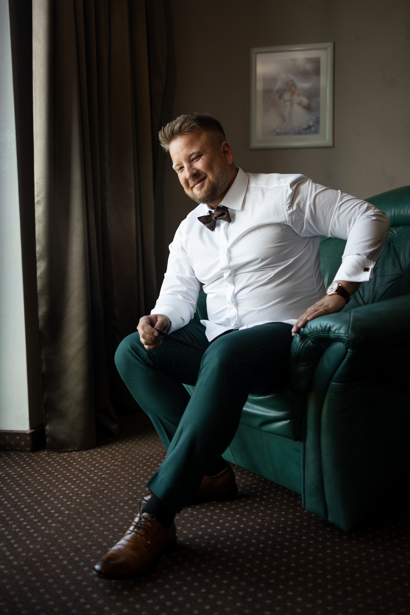 Elegancki mężczyzna w białej koszuli i zielonych spodniach siedzi w fotelu przy oknie, z brązową muchą i skórzanym paskiem zegarka.