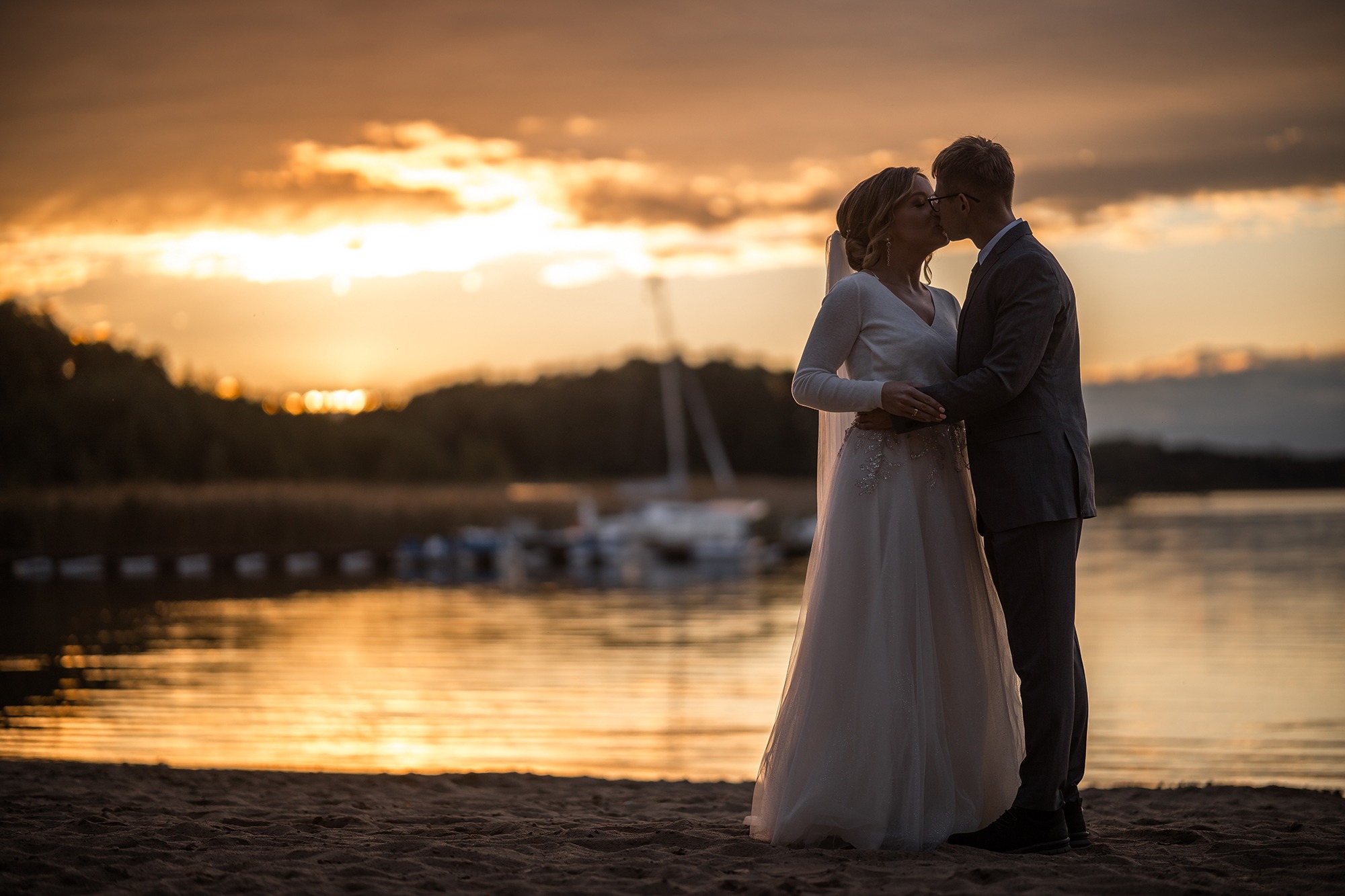 Para młoda całuje się na plaży o zachodzie słońca, w tle woda i łodzie.