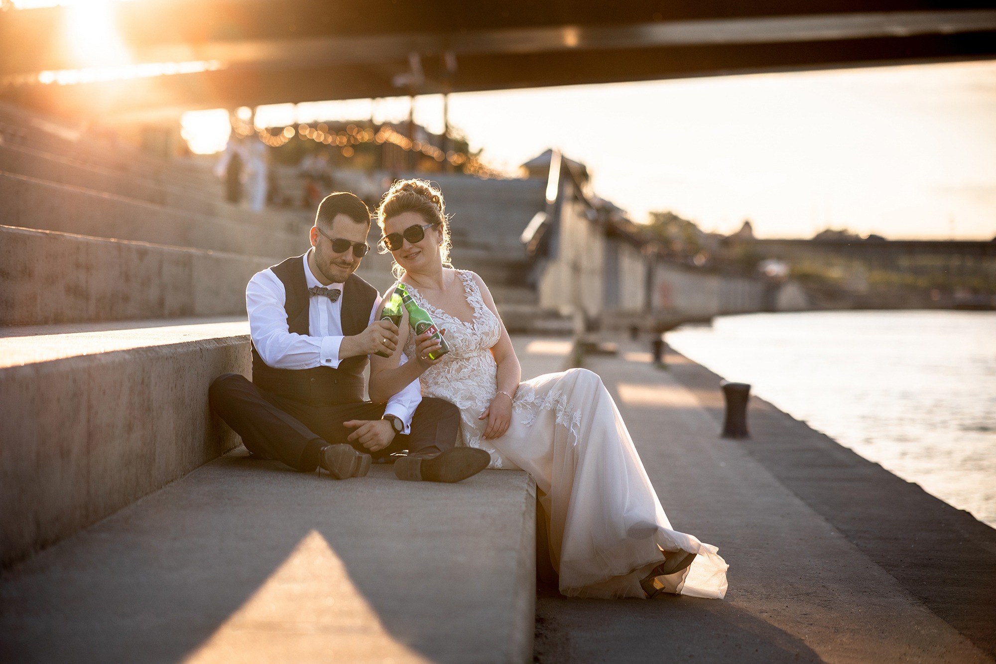 Para młoda w okularach przeciwsłonecznych siedzi na schodach nad Wisłą w Warszawie, pijąc piwo w zielonych butelkach. W tle widoczne oświetlenie i elementy architektury.