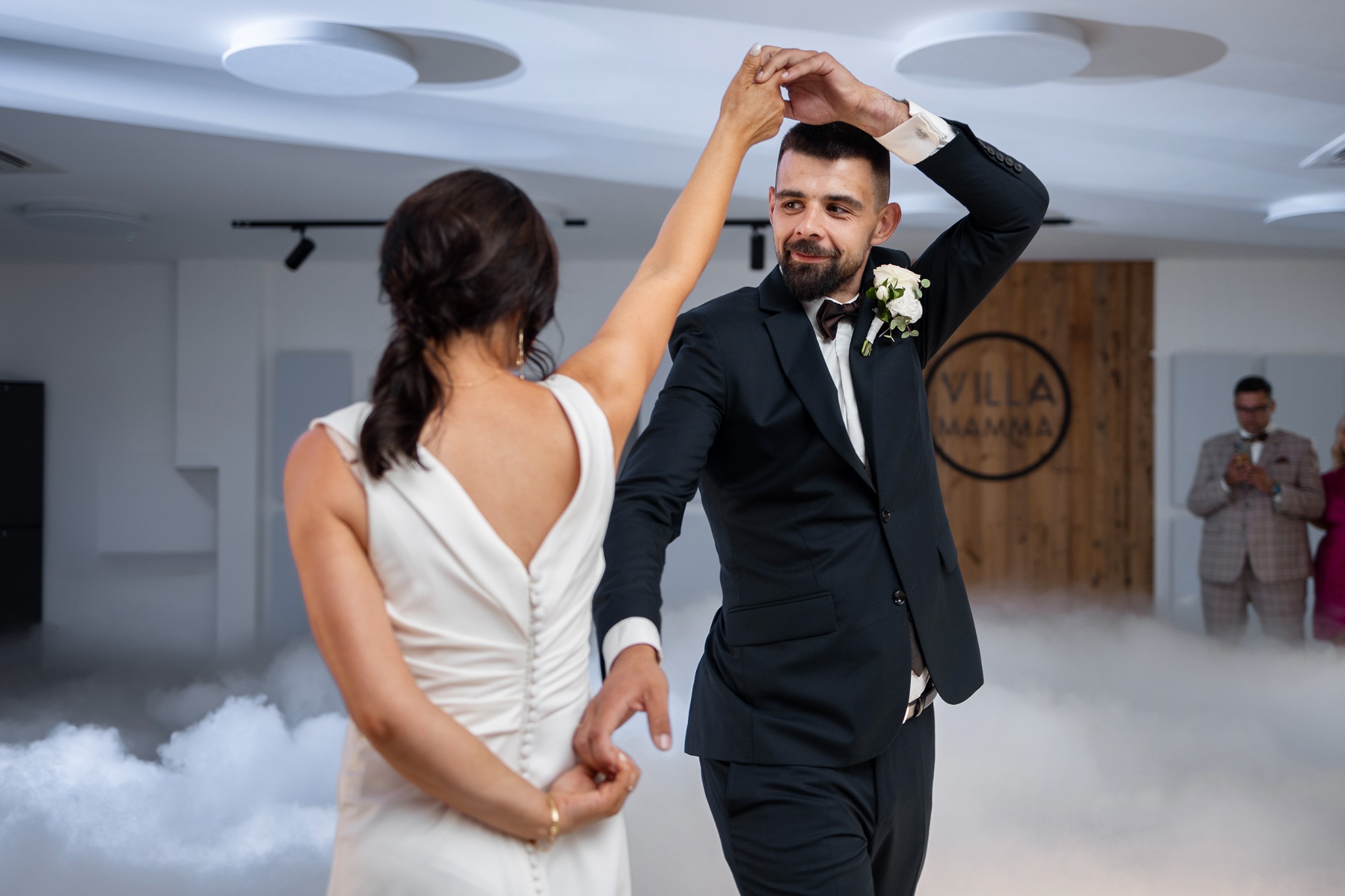 Para młoda tańczy w dymie na sali weselnej, pan młody w czarnym garniturze z butonierką, panna młoda w białej sukni z odkrytymi plecami.