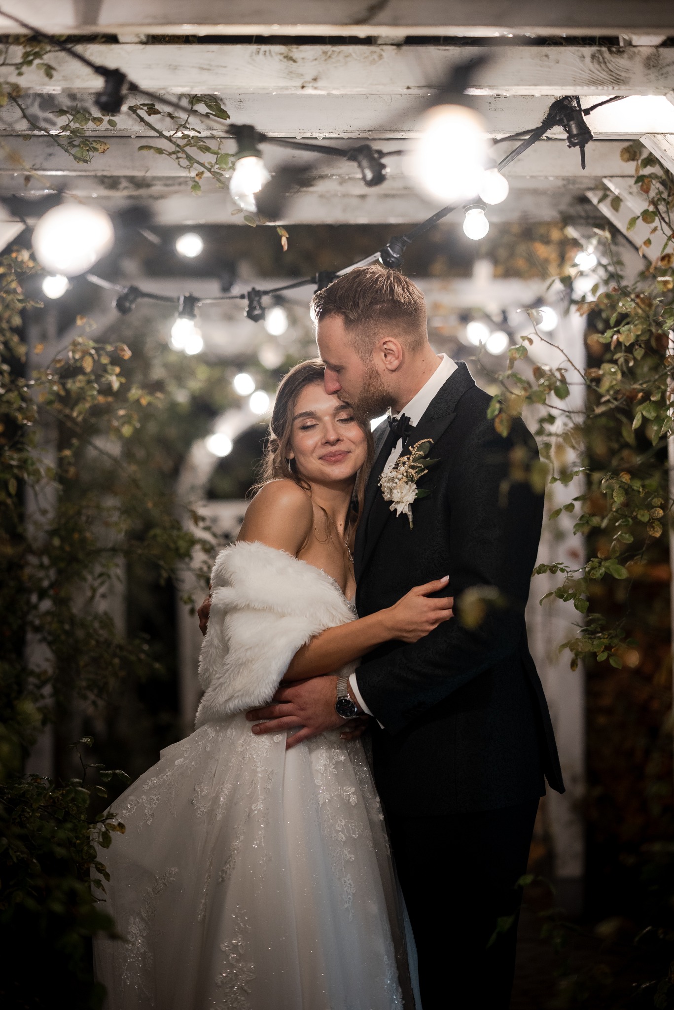 Elegancka para młoda w objęciach pod drewnianą pergolą z girlandami świetlnymi. Pan młody całuje pannę młodą w czoło. Romantyczna atmosfera.