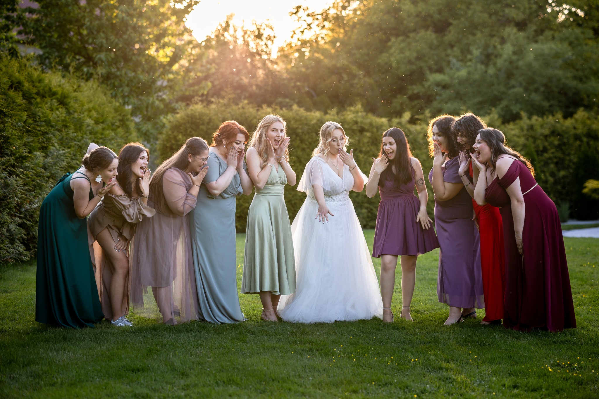 Panna młoda w białej sukni i druhny w kolorowych sukniach, stojące na trawie z zaskoczonymi minami na tle zieleni i zachodzącego słońca. Ujęcie z plenerowej sesji ślubnej.