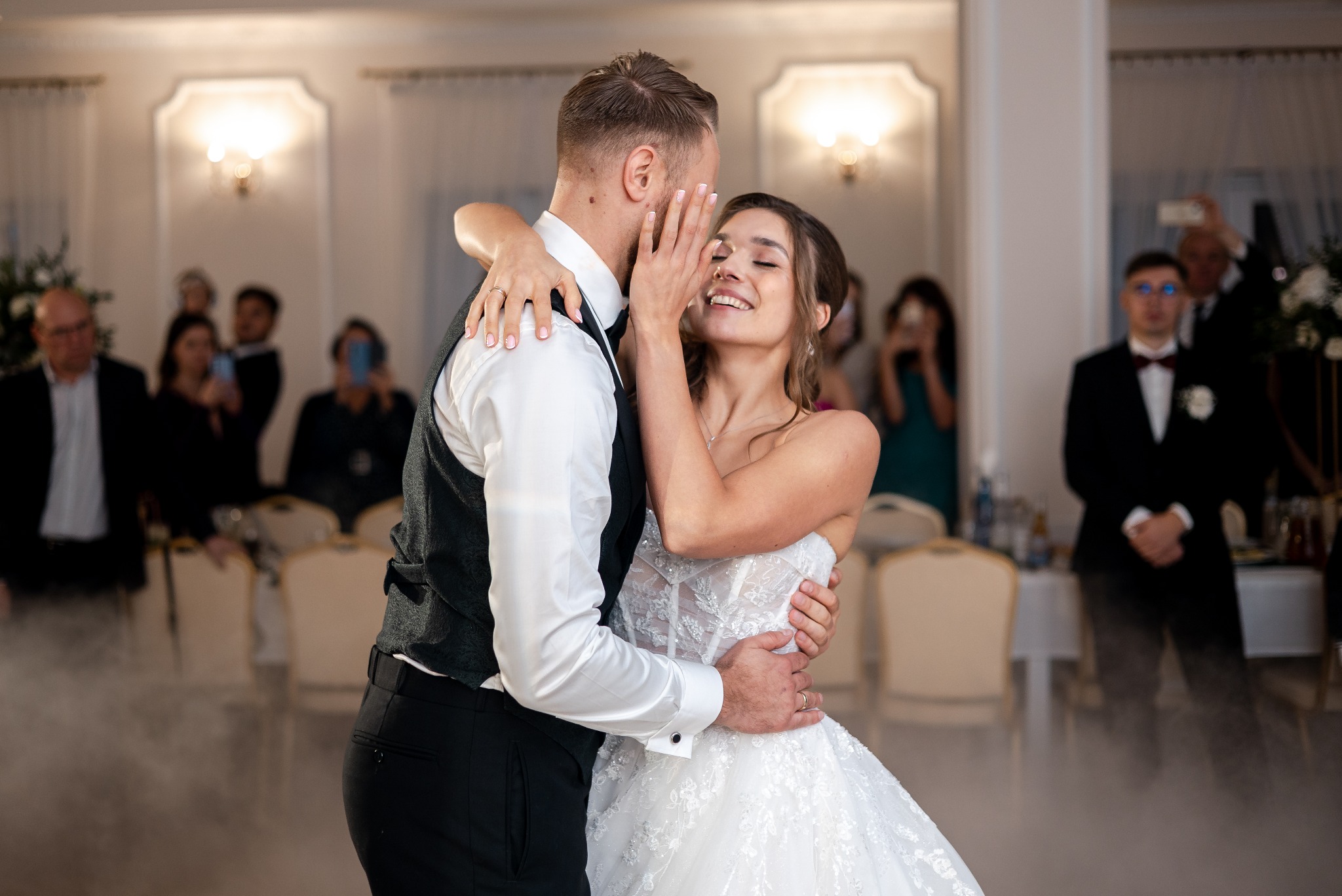 Uśmiechnięta panna młoda w objęciach pana młodego podczas pierwszego tańca, w tle goście weselni i dekoracje sali.