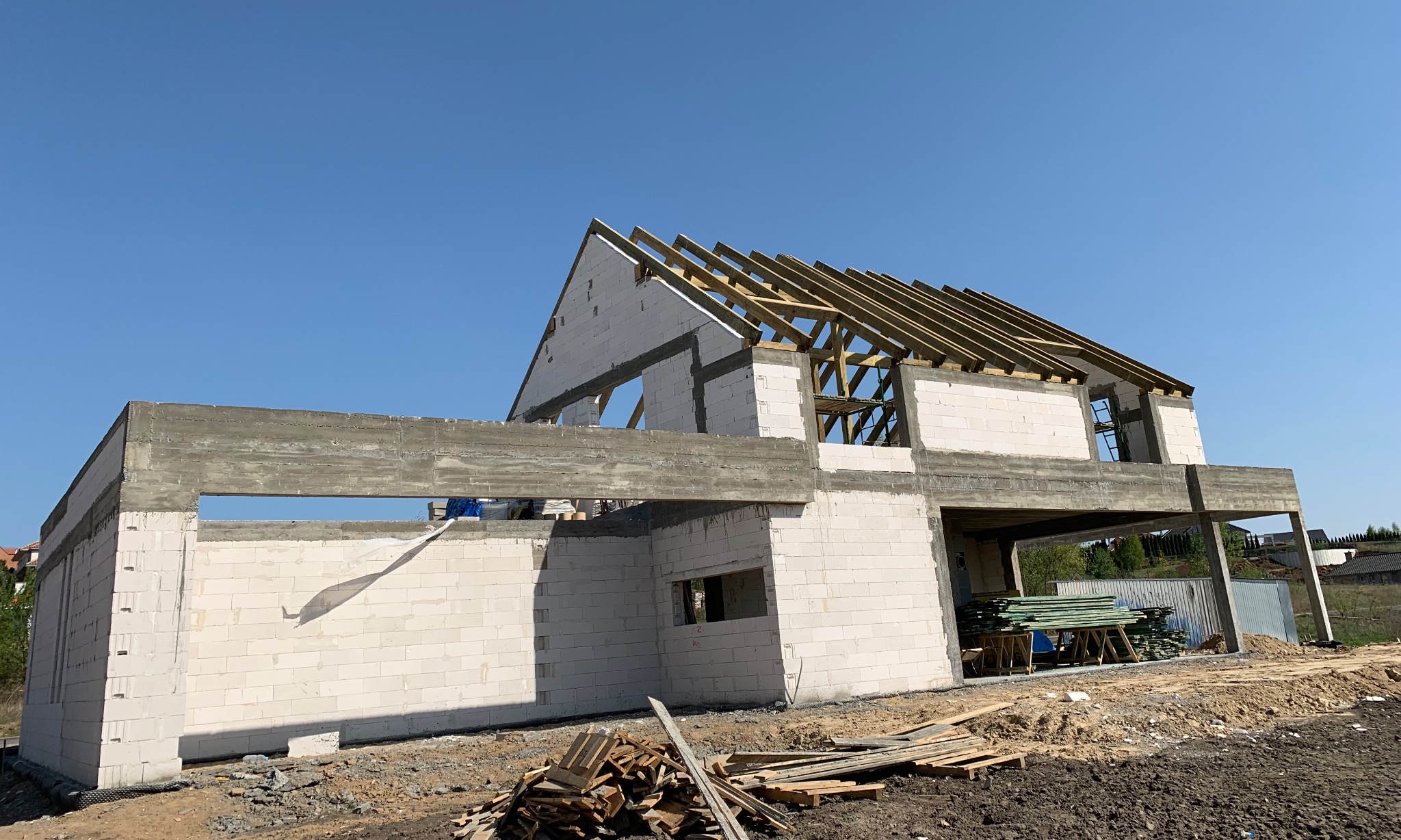 Surowy stan domu jednorodzinnego w trakcie budowy: bloczki z betonu komórkowego, betonowe nadproża, drewniana konstrukcja dachu w słoneczny dzień.