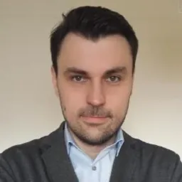 Portret mężczyzny w ciemnej marynarce i jasnej koszuli, z krótką brodą i ciemnymi włosami, na jasnym tle.