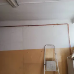 Miedziana rura centralnego ogrzewania zamontowana wzdłuż ściany nad grzejnikiem, widoczna drabina malarska.