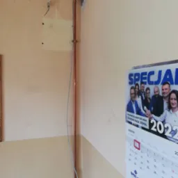 Miedziana rura pionowa zamontowana przy ścianie, widoczne uchwyty mocujące, ściana w kolorze beżowym, obok wisi kalendarz.