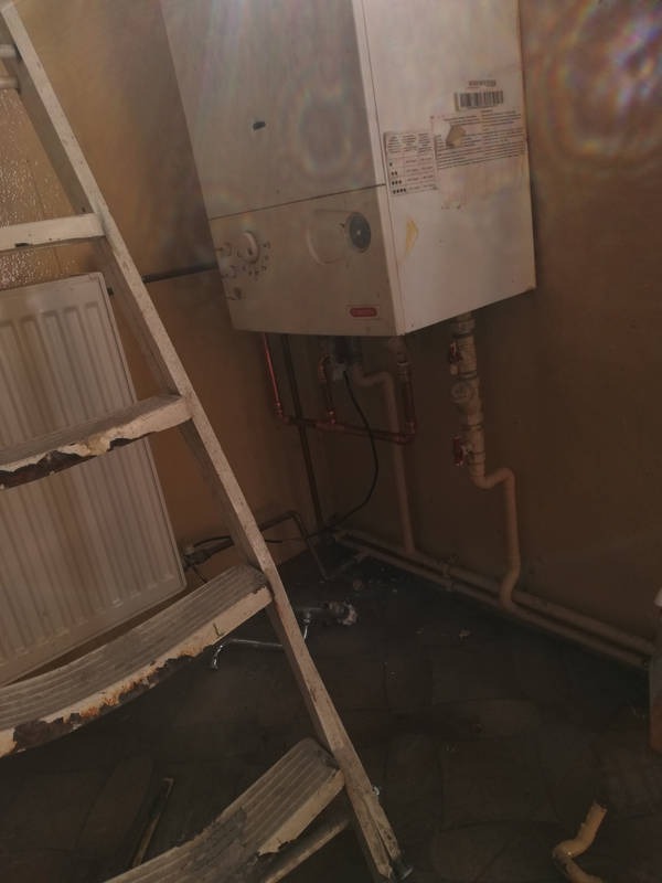 Biały, wiszący piec gazowy z widocznymi miedzianymi rurami, zamontowany w pomieszczeniu z żółtymi ścianami, obok drabina malarska i grzejnik.