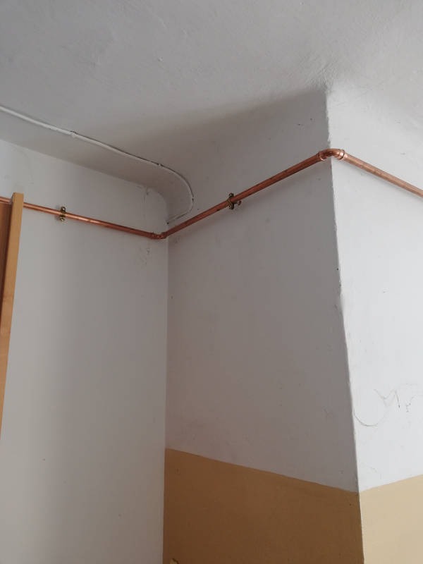 Miedziane rury centralnego ogrzewania zamocowane do ściany i sufitu, widoczne obejmy mocujące i kolanko, ściany pomalowane farbą w dwóch kolorach.