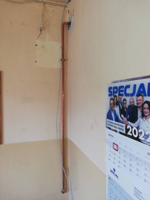 Miedziana rura pionowa zamontowana przy ścianie, widoczne uchwyty mocujące, ściana w kolorze beżowym, obok wisi kalendarz.