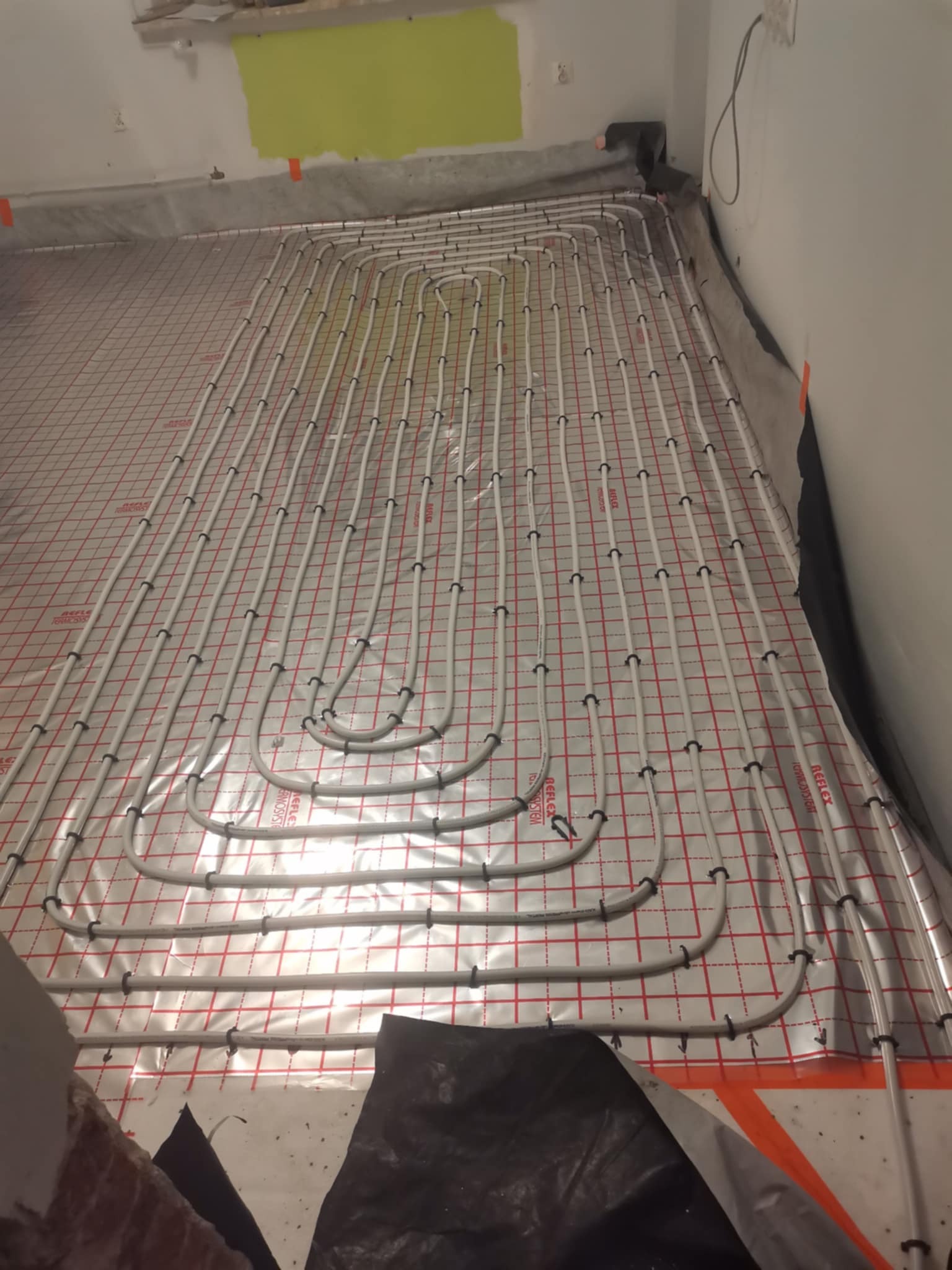 Ułożone rury ogrzewania podłogowego na folii z czerwoną siatką, widoczne czarne uchwyty mocujące, fragment ściany z zielonym śladem farby.