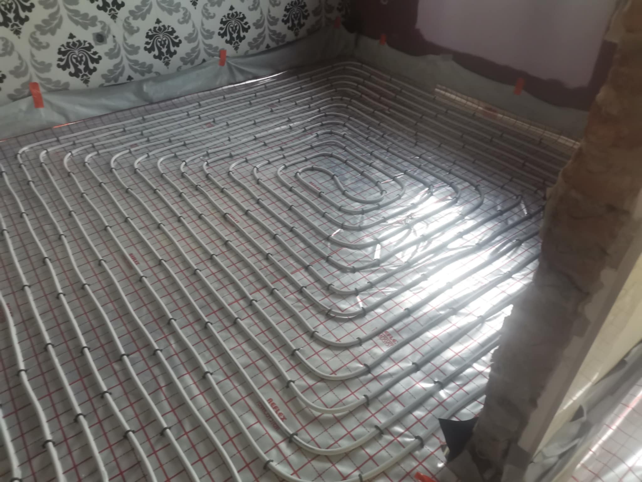 Białe rury ogrzewania podłogowego ułożone na folii z czerwoną siatką w pomieszczeniu w Parysowie. Widoczne fragmenty ścian i podłoża.