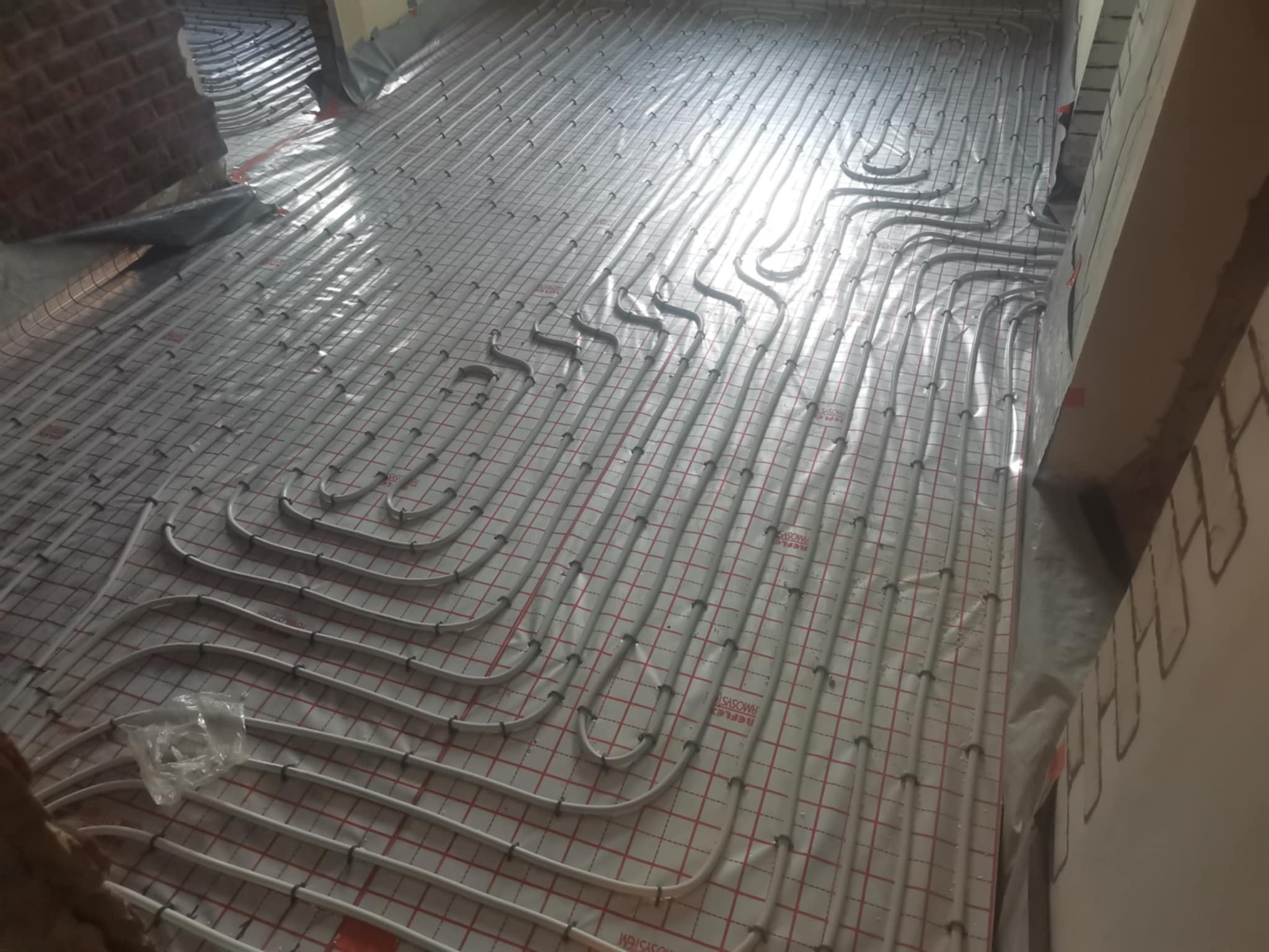 Białe rury ogrzewania podłogowego ułożone równolegle na folii z czerwoną siatką, gotowe do zalania posadzką. Widok z góry, z bliska.