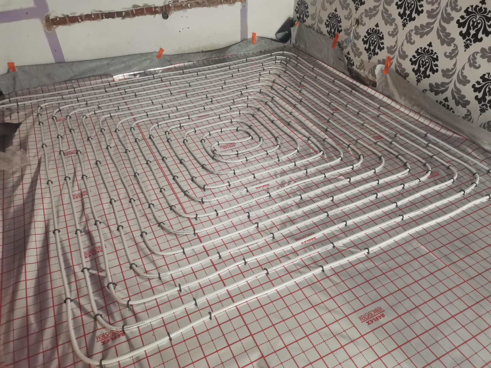 Białe rury ogrzewania podłogowego ułożone spiralnie na folii z czerwoną kratką, widoczne fragmenty ścian w trakcie wykończenia.