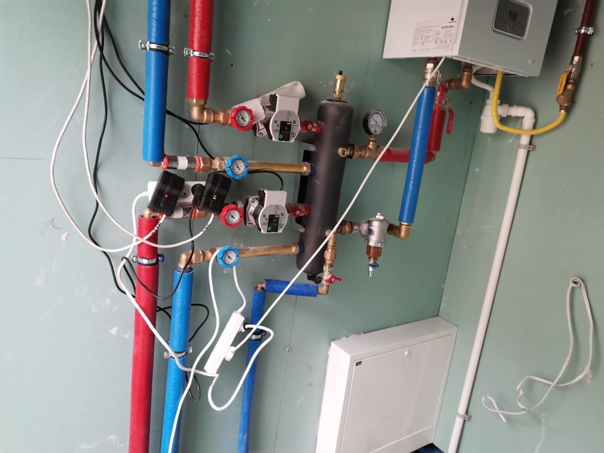 Instalacja grzewcza: kotłownia z rurami w kolorach czerwonym i niebieskim, manometry, pompy obiegowe i biały zasobnik. Widoczne przewody elektryczne.