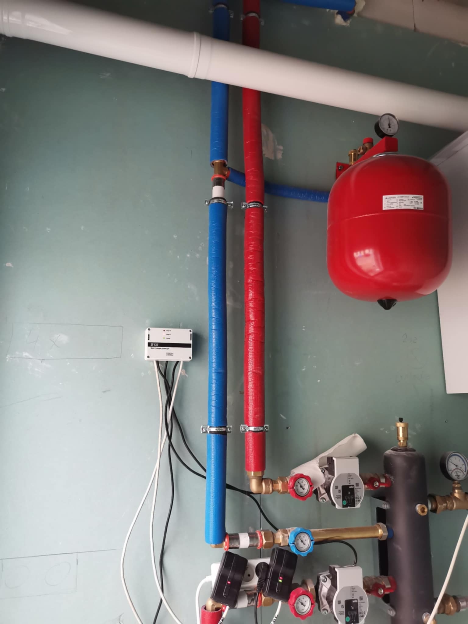 Instalacja grzewcza z czerwonym zbiornikiem wyrównawczym, rurami w izolacji (czerwona i niebieska) oraz białym sterownikiem na ścianie w surowym stanie.