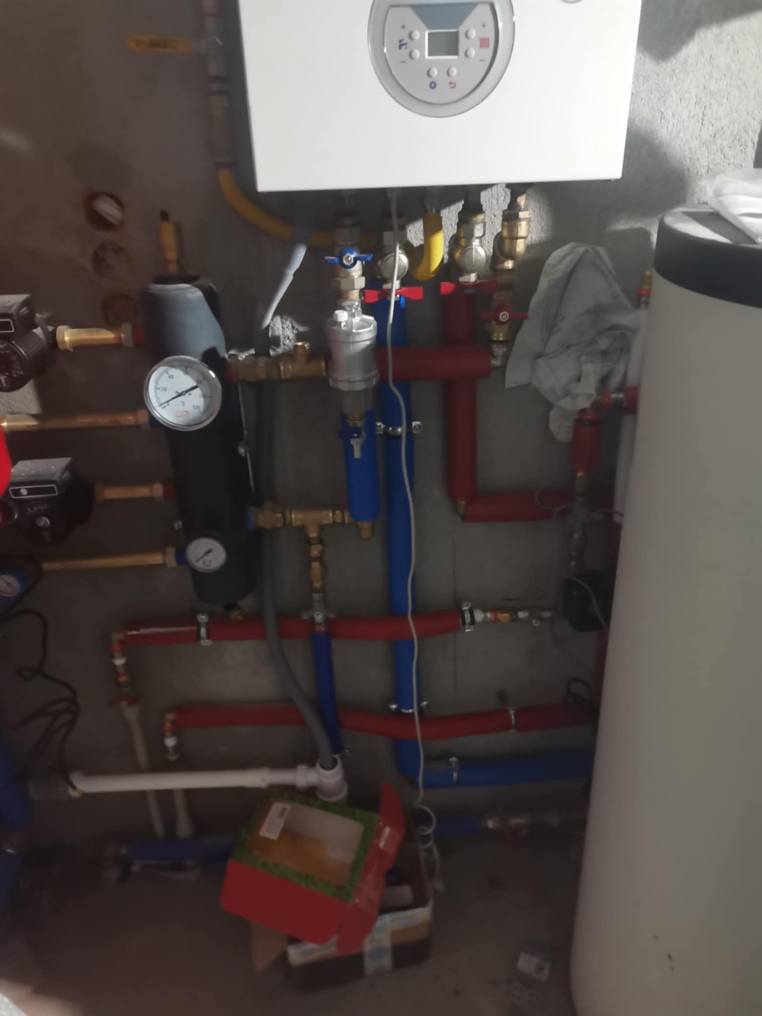 Biały piec gazowy z podłączeniami rur miedzianych, plastikowych (czerwonych i niebieskich) oraz zaworów. Widoczne manometry i elementy instalacji grzewczej.