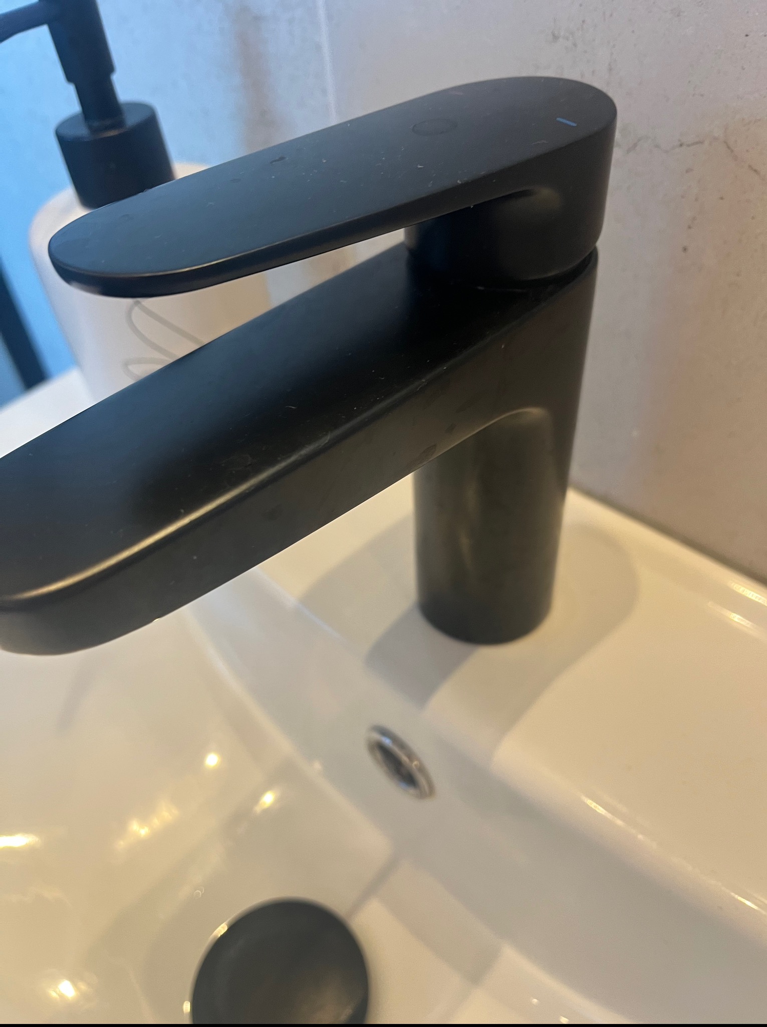 Nowoczesna, matowa, czarna bateria umywalkowa zamontowana na białej umywalce. W tle dozownik mydła i beżowa ściana. Minimalistyczny design w łazience.